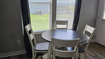 Un comedor con una mesa redonda y cuatro sillas, ventanas amplias con persianas, una pared gris y suelo de madera.