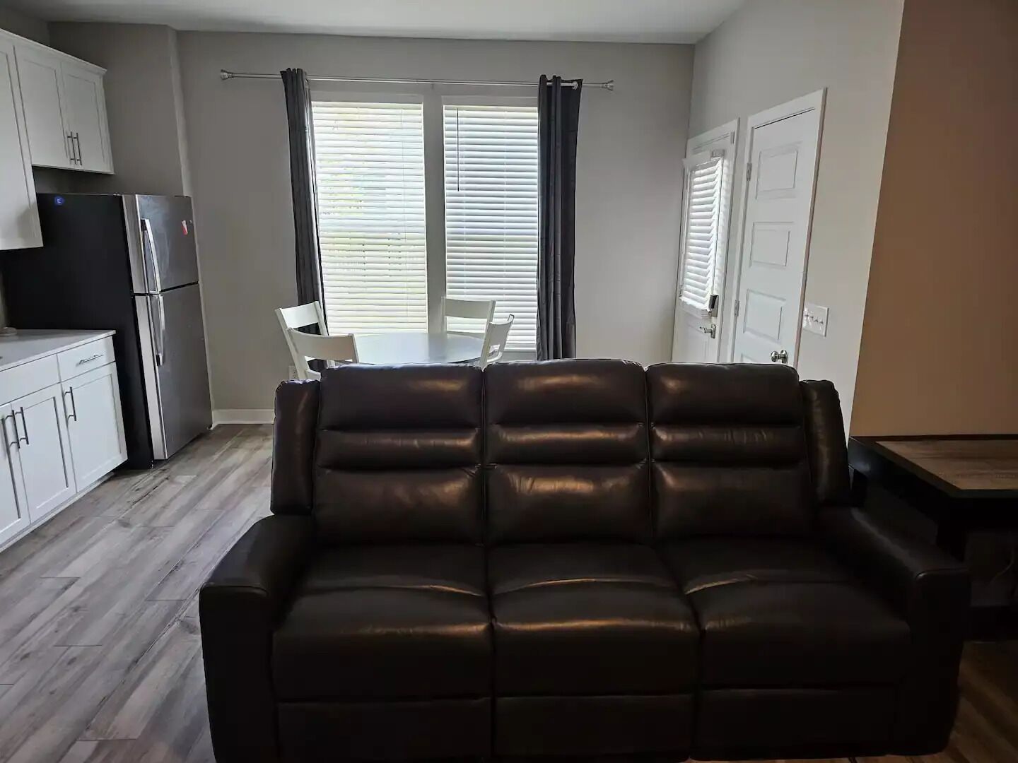 A living room with a dark leather sofa, a dining table with chairs, a kitchen area with cabinets and a refrigerator, and a window with blinds.