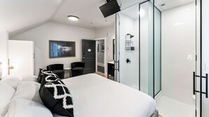 Quarto moderno com cama grande, box de vidro, TV na parede e um pequeno espaço de estar com cadeiras.
