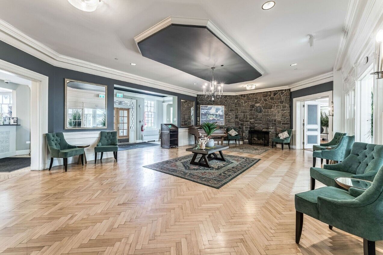 Un salon spacieux doté d’un parquet en bois disposé en point de Hongrie, de fauteuils recouverts d’un tissu vert, d’une cheminée en pierre et d’un grand miroir mural.