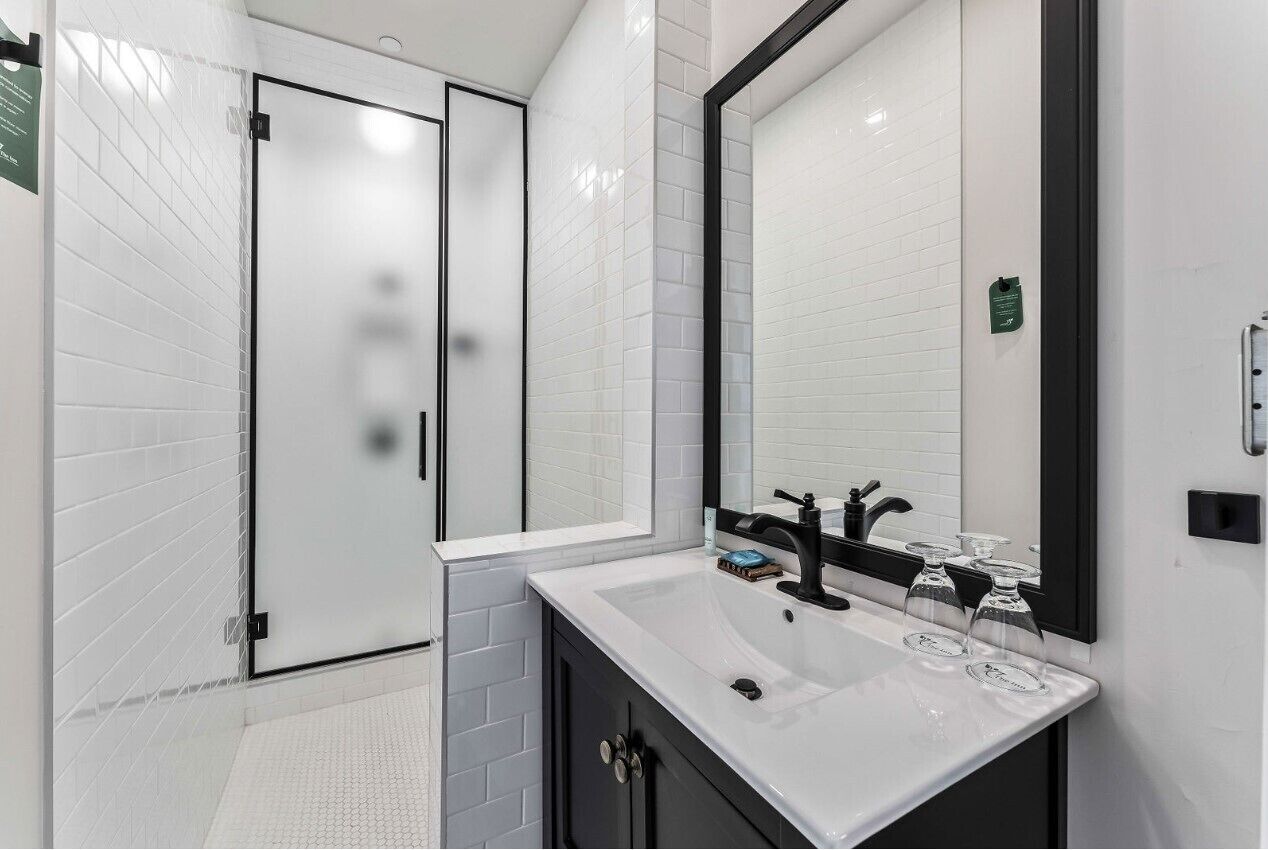 A modern bathroom with a white tiled shower area, a large mirror, and a white sink with black fixtures.
