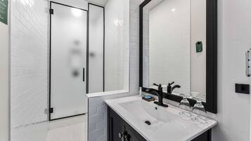 A modern bathroom with a white tiled shower area, a large mirror, and a white sink with black fixtures.