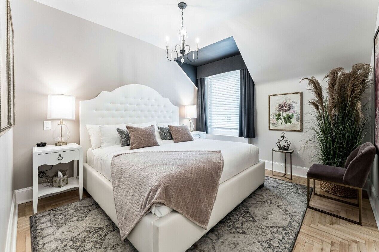 Un dormitorio con cabecera capitonada, una cama grande, una mesita de noche con lámpara, una silla y una ventana con cortinas.