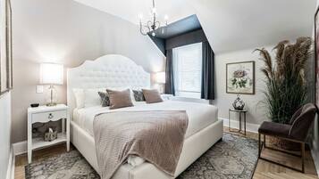 Un dormitorio con cabecera capitonada, una cama grande, una mesita de noche con lámpara, una silla y una ventana con cortinas.