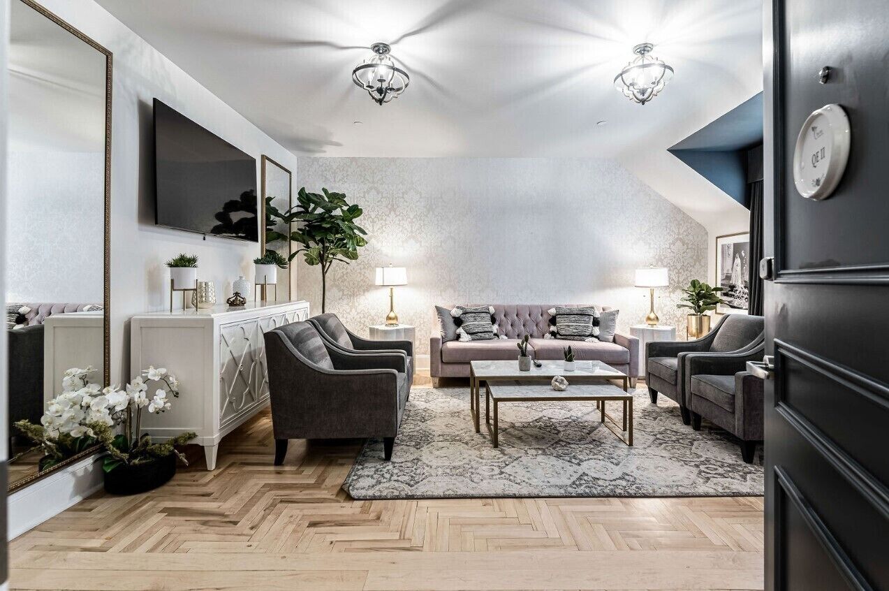 A modern living room with a sofa set, two armchairs, a coffee table, and a TV mounted on the wall. There are two side tables with lamps, a large mirror, and a potted plant.