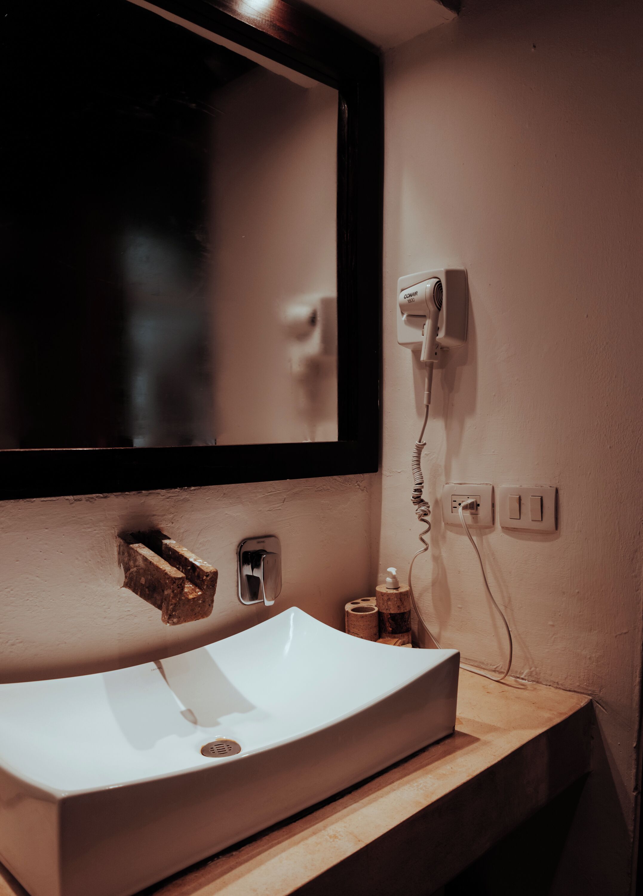 Une salle de bain avec un lavabo blanc, un miroir, un sèche-cheveux et un distributeur de savon.