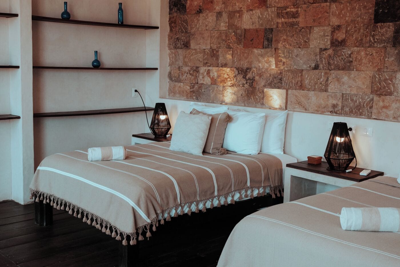 A hotel room with two beds, a stone wall, and a shelf with decorative items.