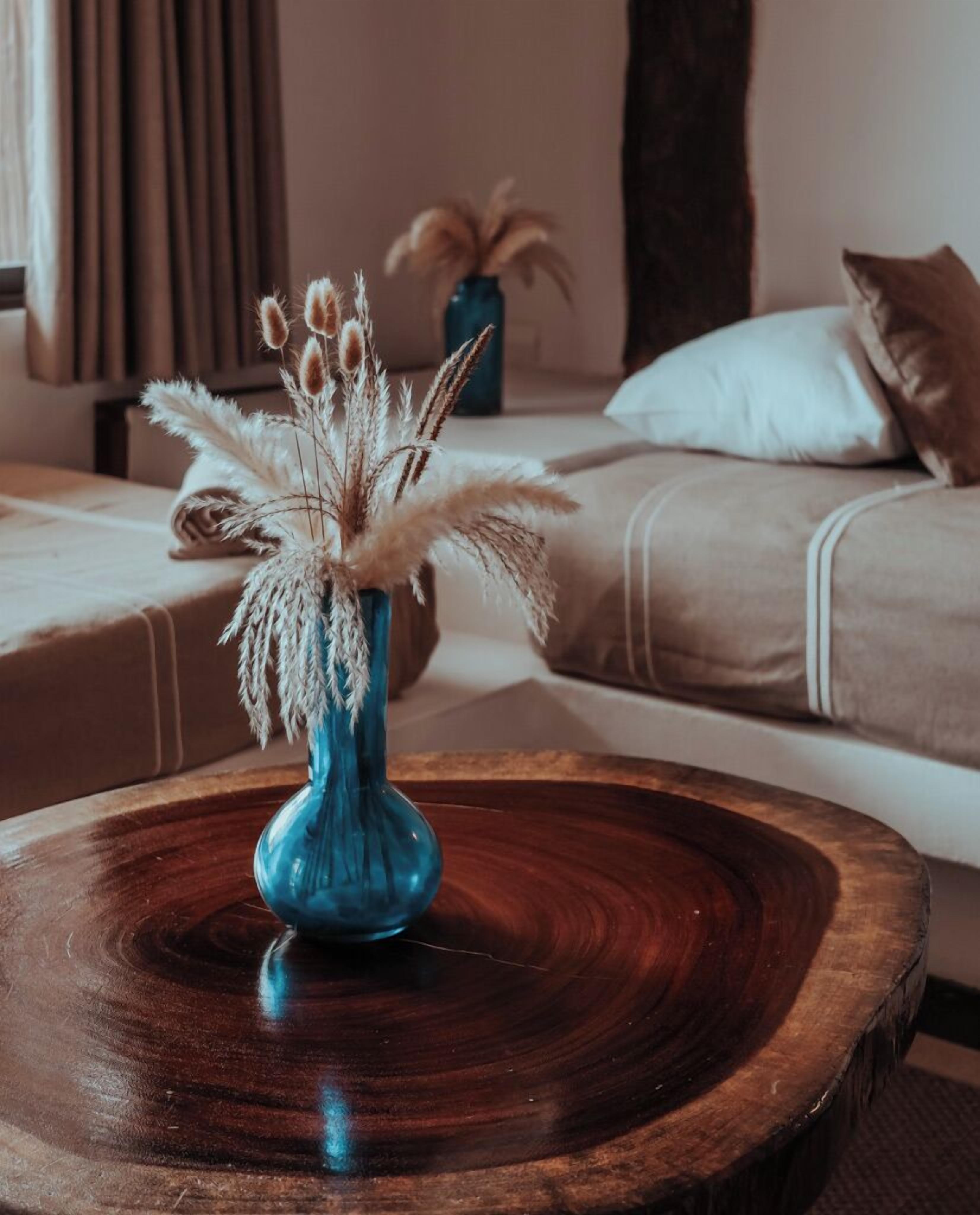 A room with a wooden coffee table in the foreground, a blue vase with dried plants, and a sofa with cushions in the background.