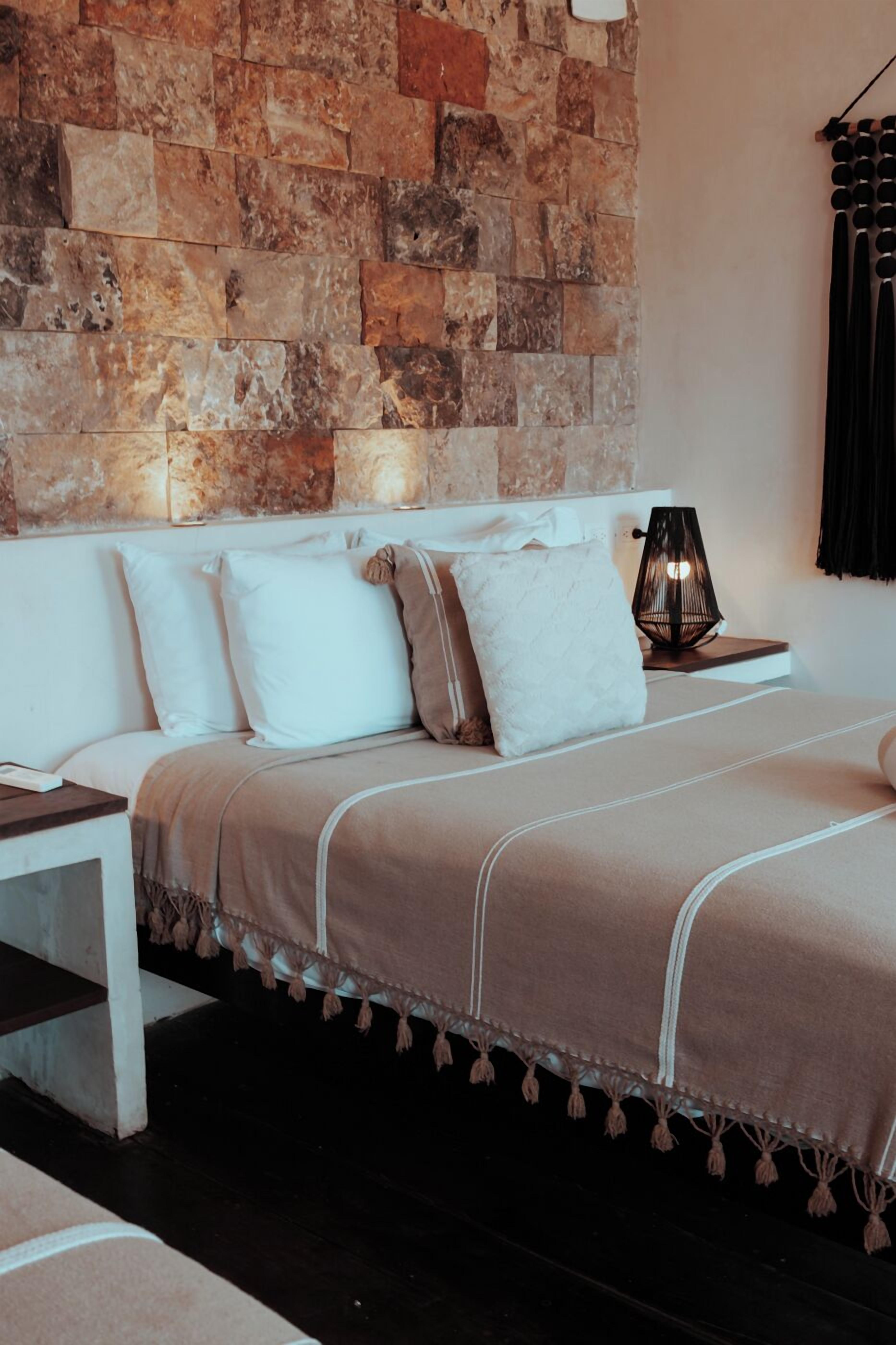 Un dormitorio con pared de piedra, cama con ropa de cama blanca y beige, mesita de noche y lámpara.