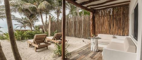 A wooden deck with a white sofa and two wooden chairs, overlooking a sandy beach with palm trees and the ocean.