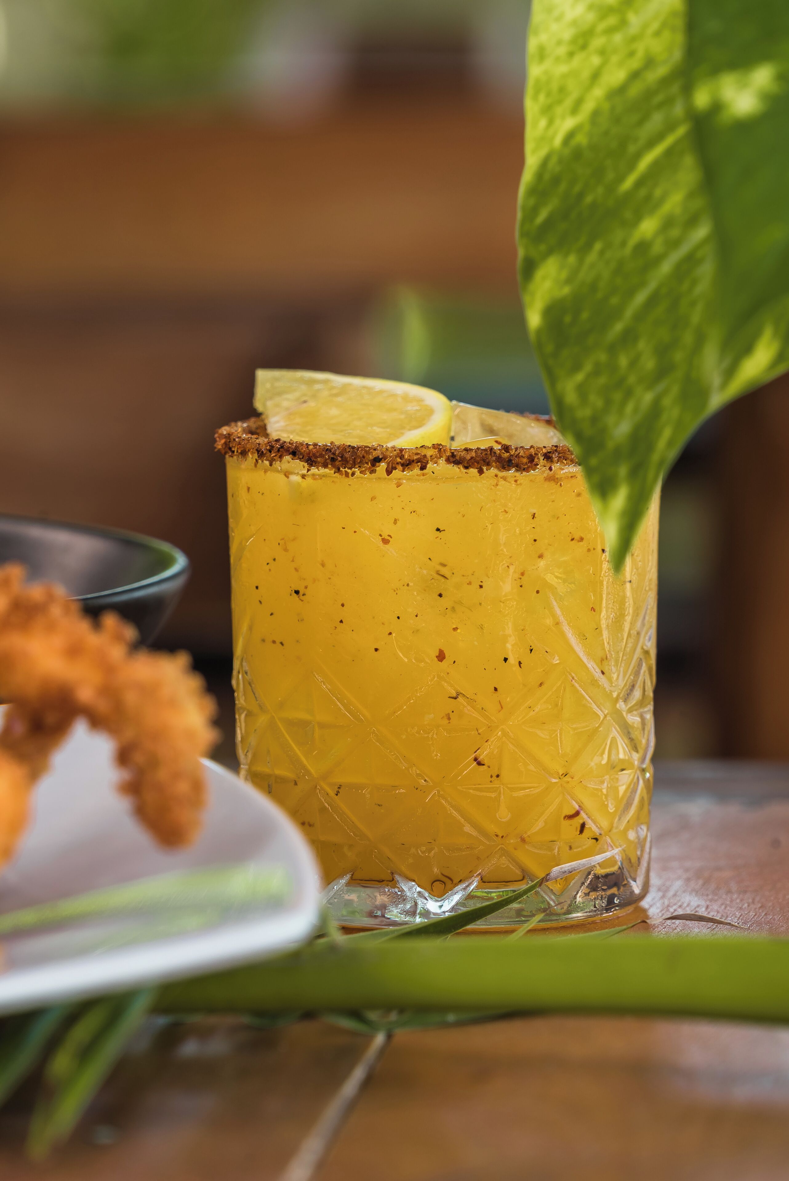 A glass of yellow drink with a lemon slice garnish, placed on a table with a green leaf.