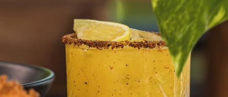 A glass of yellow drink with a lemon slice garnish, placed on a table with a green leaf.