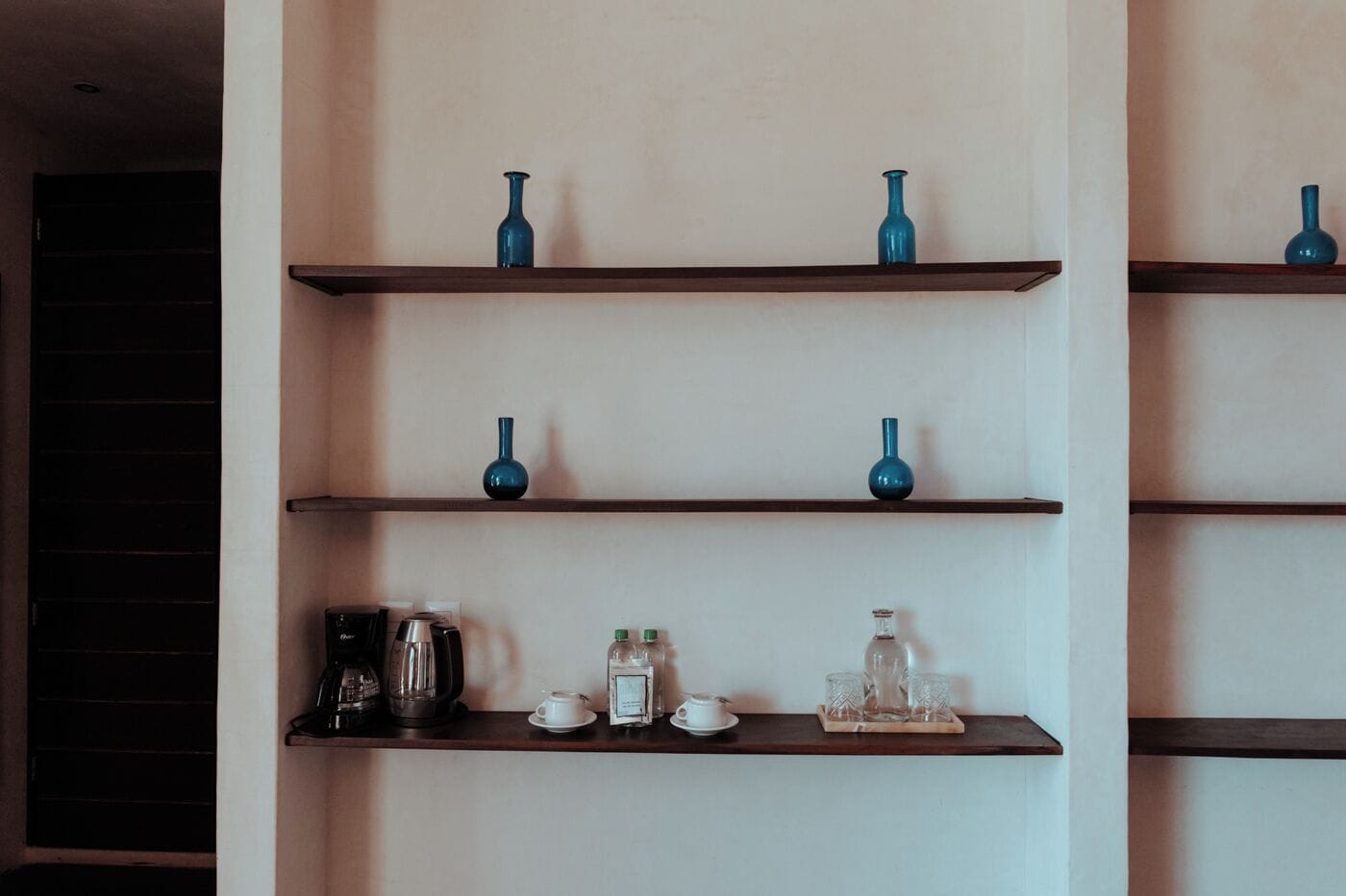 A wall with three shelves, each holding decorative vases and a coffee maker on the bottom shelf.