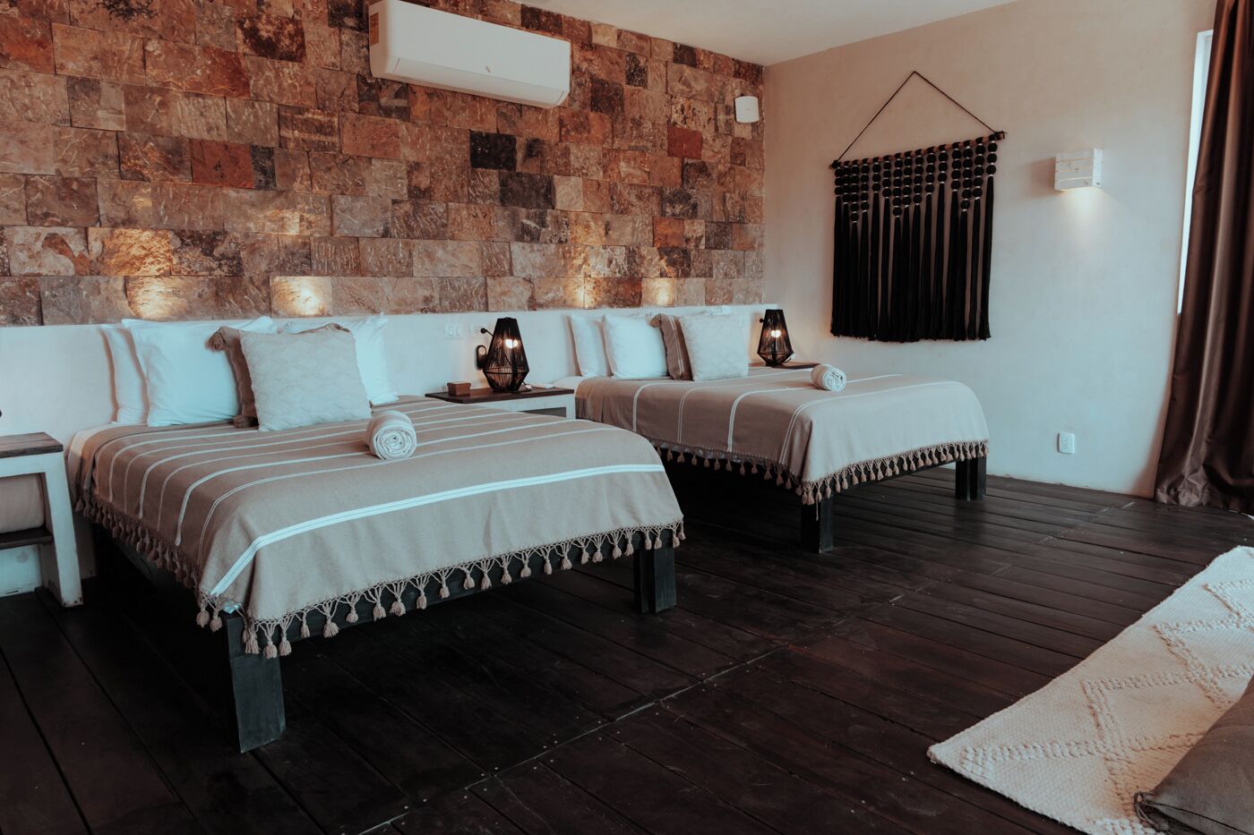 A bedroom with two beds, a stone wall, a window with curtains, and a wooden floor.