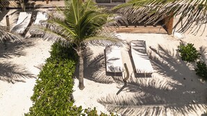 Dos cabañas de playa con techos de paja, barandillas de madera, paredes blancas, una playa arenosa con palmeras y sillas de descanso.