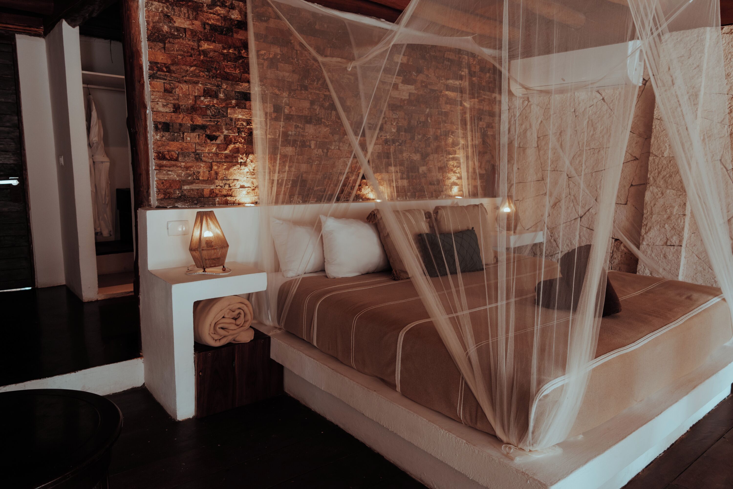 Una cama con dosel, una mesita de noche con lámpara y una pared de ladrillo al fondo.
