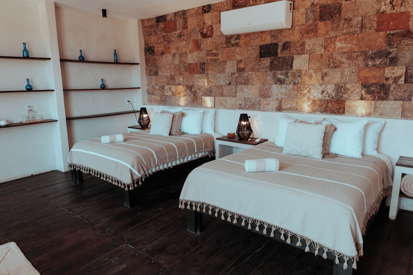 A bedroom with two beds, wooden flooring, a stone wall, and a white air conditioning unit.