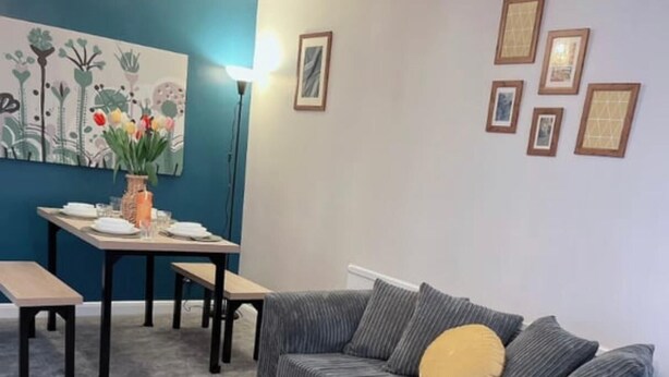 Ein Wohnzimmer mit grauem Sofa, einem Holztisch, einem Esstisch mit Stühlen und einem Gemälde an der Wand.