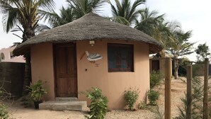 A small, single-story building with a thatched roof, a wooden door, and a window with a red frame. - Case Typique Sénégalaise Piscine vue mer (Palmarin)