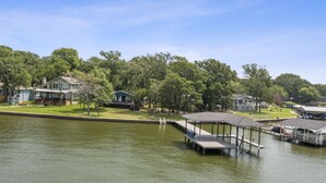A lakeside property with a dock, a covered boat launch, and a grassy area with houses in the background. - Open Water~Sunsets~Swimming~Standing Work Station with High Speed Internet~Lake (Mabank)