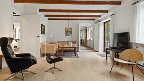 En rummelig stue med højt til loftet, træbjælker, en sofa, et sofabord, et fjernsyn og et spiseområde med bord og stole.