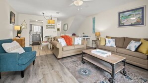 Una sala de estar moderna con un sillón azul, sofás beige y una mesa de centro. Hay un comedor con una mesa y sillas, y una cocina al fondo.