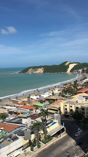 A coastal town with buildings along the beach, a clear sky, and a hilly landscape in the distance.