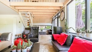 Amplio salón tipo loft con techo alto, una escalera, un sofá con cojines rojos, una mesa de centro con cactus en macetas y un gran ventanal con un reloj.