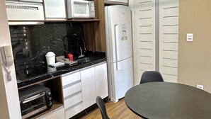 A compact kitchen with white cabinets, a microwave, a refrigerator, a toaster, and a round table with chairs. - Saint Sebastian 307 Duplex no Centro. (Jaragua do Sul)