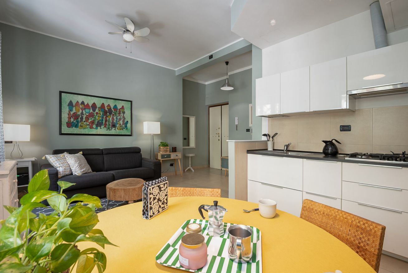 Una sala de estar moderna con un sofá, una mesa de comedor con una bandeja de objetos, una cocina con armarios blancos y un ventilador de techo.