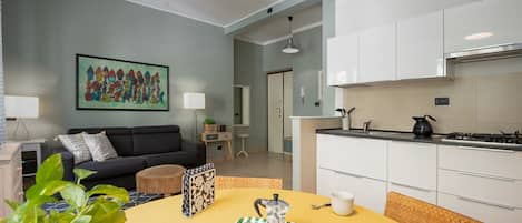 A modern living room with a sofa, a dining table with a tray of items, a kitchen with white cabinets, and a ceiling fan.