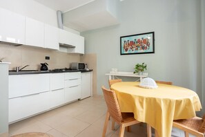 Une cuisine moderne avec des meubles blancs, une table à manger recouverte d’une nappe jaune, des chaises en osier et une œuvre d’art encadrée accrochée au mur.