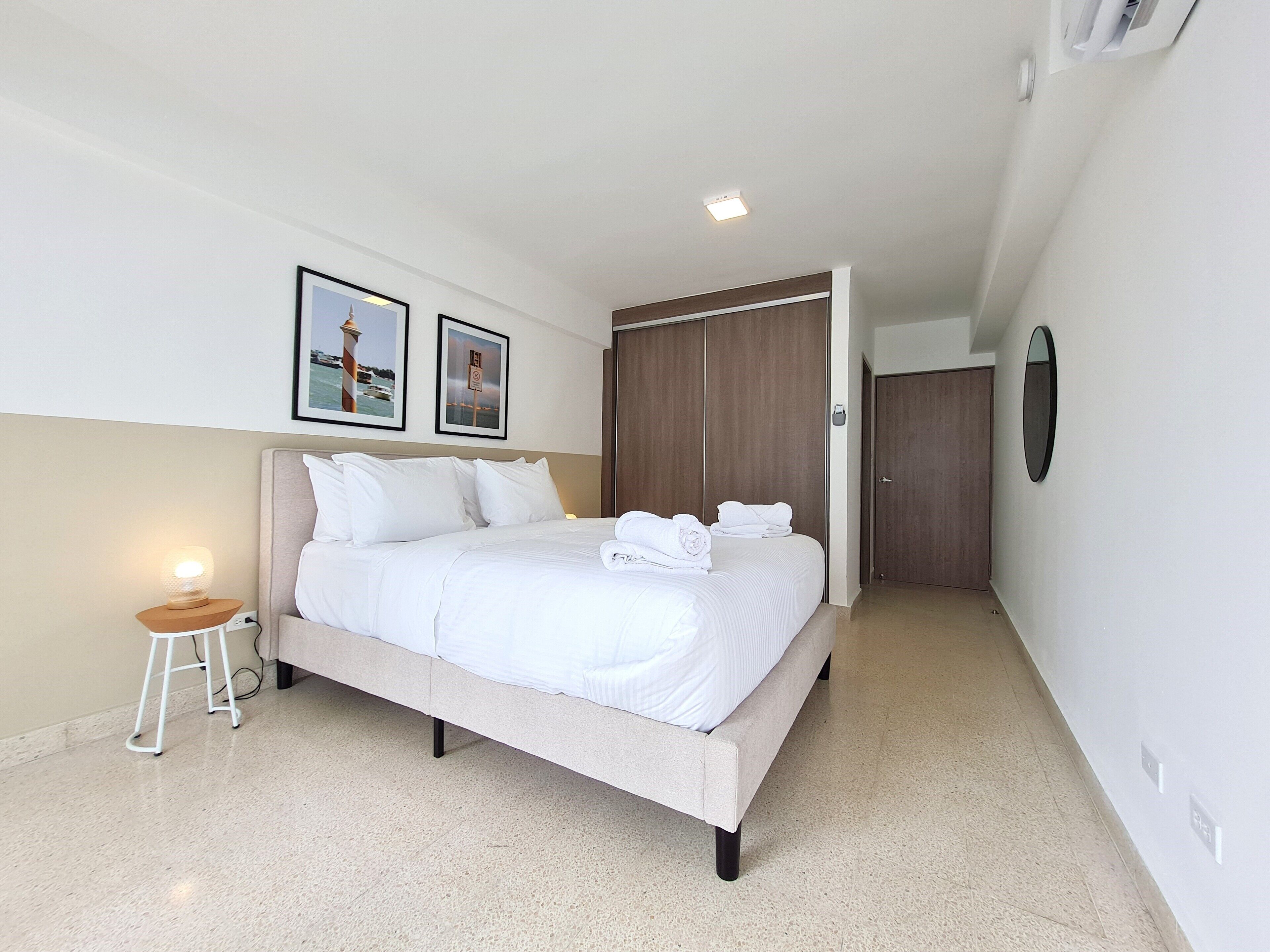 A neatly made bed with white linens, a small bedside table with a lamp, and two framed pictures on the wall.