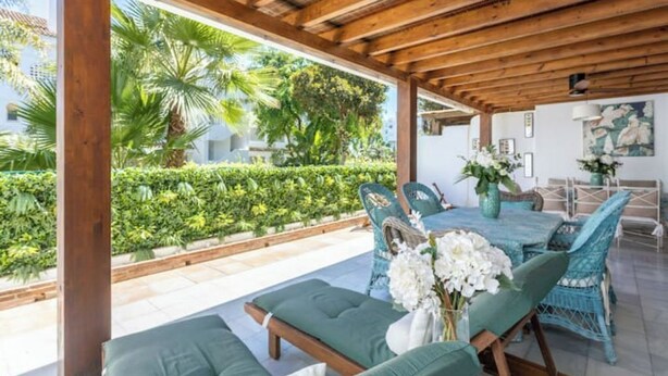 A covered outdoor patio with wooden beams, a table set with chairs, and a view of a well-maintained garden. - Magnífico Apartamento guadalmina baja  (Marbella)
