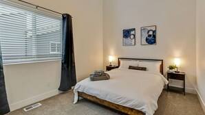 Un dormitorio con una cama, dos mesitas de noche con lámparas, una ventana con persianas y dos obras de arte colgadas en la pared.