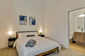 Un dormitorio con cama, dos mesitas de noche con lámparas, una mesita auxiliar, una obra de arte en la pared y un baño visible a través de una puerta abierta.