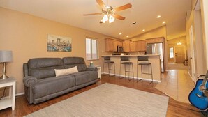 A modern living room with a gray sofa, a ceiling fan, and a kitchen area with a bar counter.