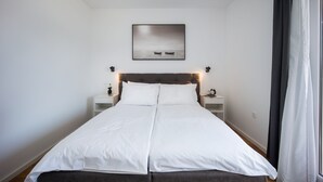 Una cama doble con ropa de cama blanca, cabecero, mesitas de noche con lámparas y un cuadro enmarcado sobre la cama.