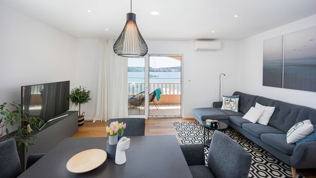 Una sala de estar moderna con televisor de pantalla plana, sofá gris, alfombra estampada y vista al mar a través de la puerta de cristal.