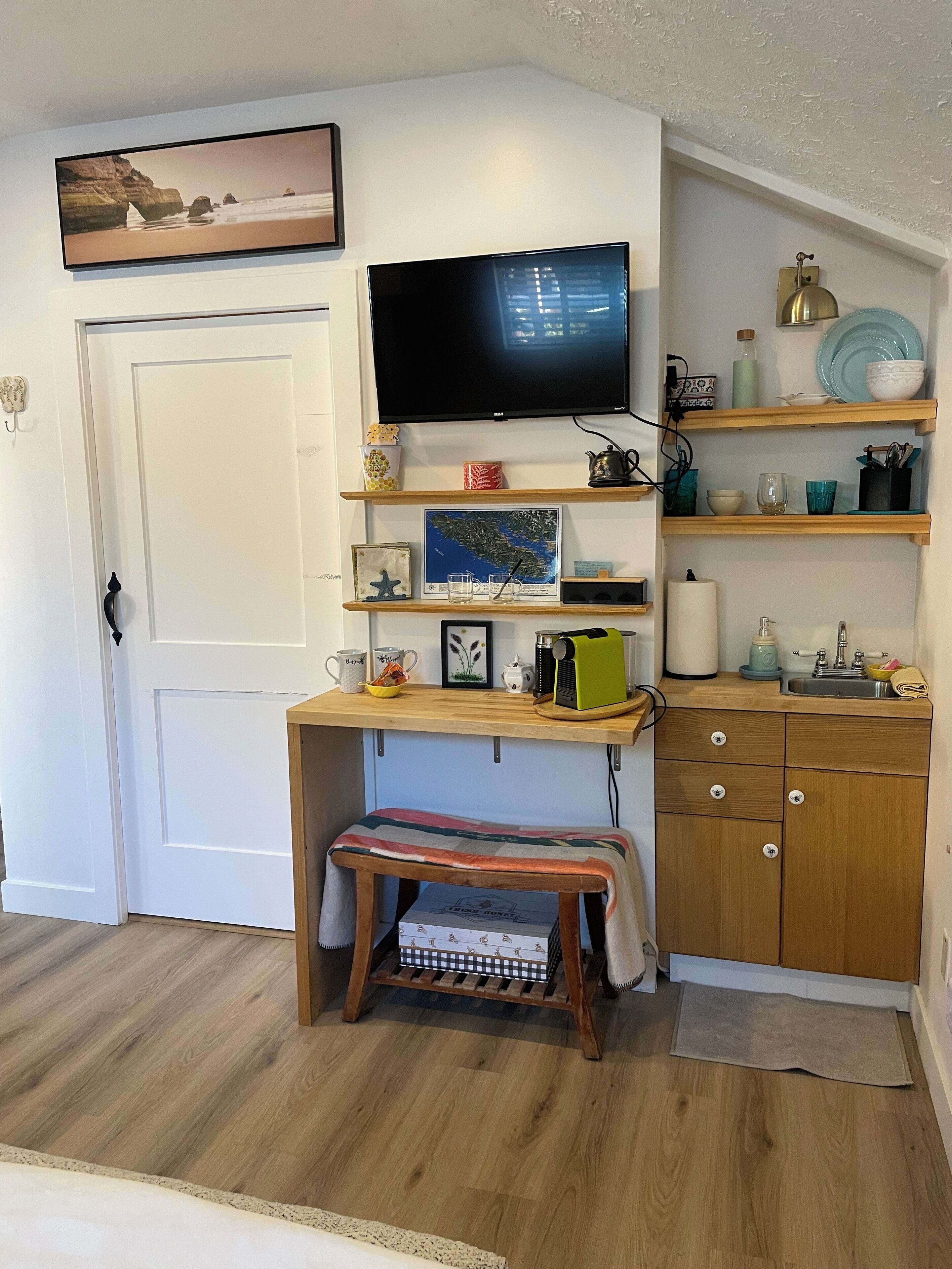 A compact living space with a wooden desk, a mounted TV, and a kitchen area with wooden cabinets and shelves.
