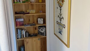 Ein Flur mit einem hölzernen Bücherregal, das Bücher, Dekoration und eine kleine Vitrine beherbergt. An der Wand hängt ein gerahmtes Bild von einer Blume, und auf dem Boden liegt eine Fußmatte mit der Aufschrift 'life is Sweet'.