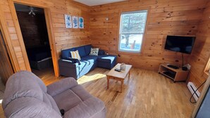 Ein gemütliches Blockhauszimmer mit Holzvertäfelung, einer Couch, einem kleinen Tisch, einem Fernseher und einem Fenster mit Blick auf Bäume.