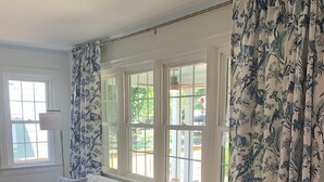 En vit soffa med gråa kuddar, ett soffbord i glas och flätade stolar i ett rum med vita väggar och blommiga gardiner.