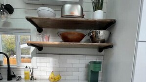 A kitchen with white cabinets, a white countertop, a hanging pot rack, a green water dispenser, and a white furry chair. - Urban Tiny Home Getaway
 (Caldwell)