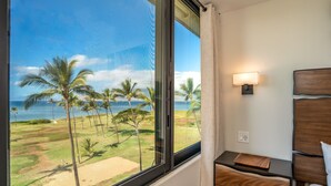 Ein Hotelzimmer mit großem Fenster, das Blick auf Palmen und einen Strand bietet. Es gibt einen Nachttisch aus Holz mit Lampe und einem kleinen Tisch.