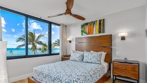 Ein Schlafzimmer mit großem Fenster und Blick auf den Ozean und Palmen, Deckenventilator, Bett mit gemusterten Bettwäsche, Nachttisch und einer Wandlampe.