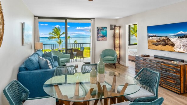 Ein modernes Wohnzimmer mit einem Glastisch, Türkis-Sofas und einem großen Fernseher, der eine Strandansicht zeigt.