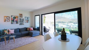 Ein modernes Wohnzimmer mit einem grauen Sofa, einem kleinen Couchtisch und einem Esstisch mit Stühlen. An den Wänden hängen abstrakte Gemälde und auf dem Esstisch steht eine Kaktee.