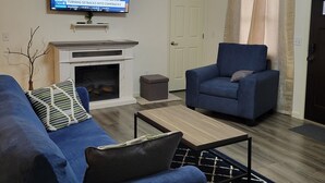 Una sala de estar con televisión, chimenea, sofá azul, mesa de centro y una puerta.