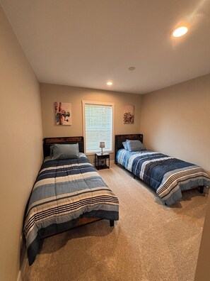 A bedroom with two beds, a window with blinds, and a small table with a lamp.